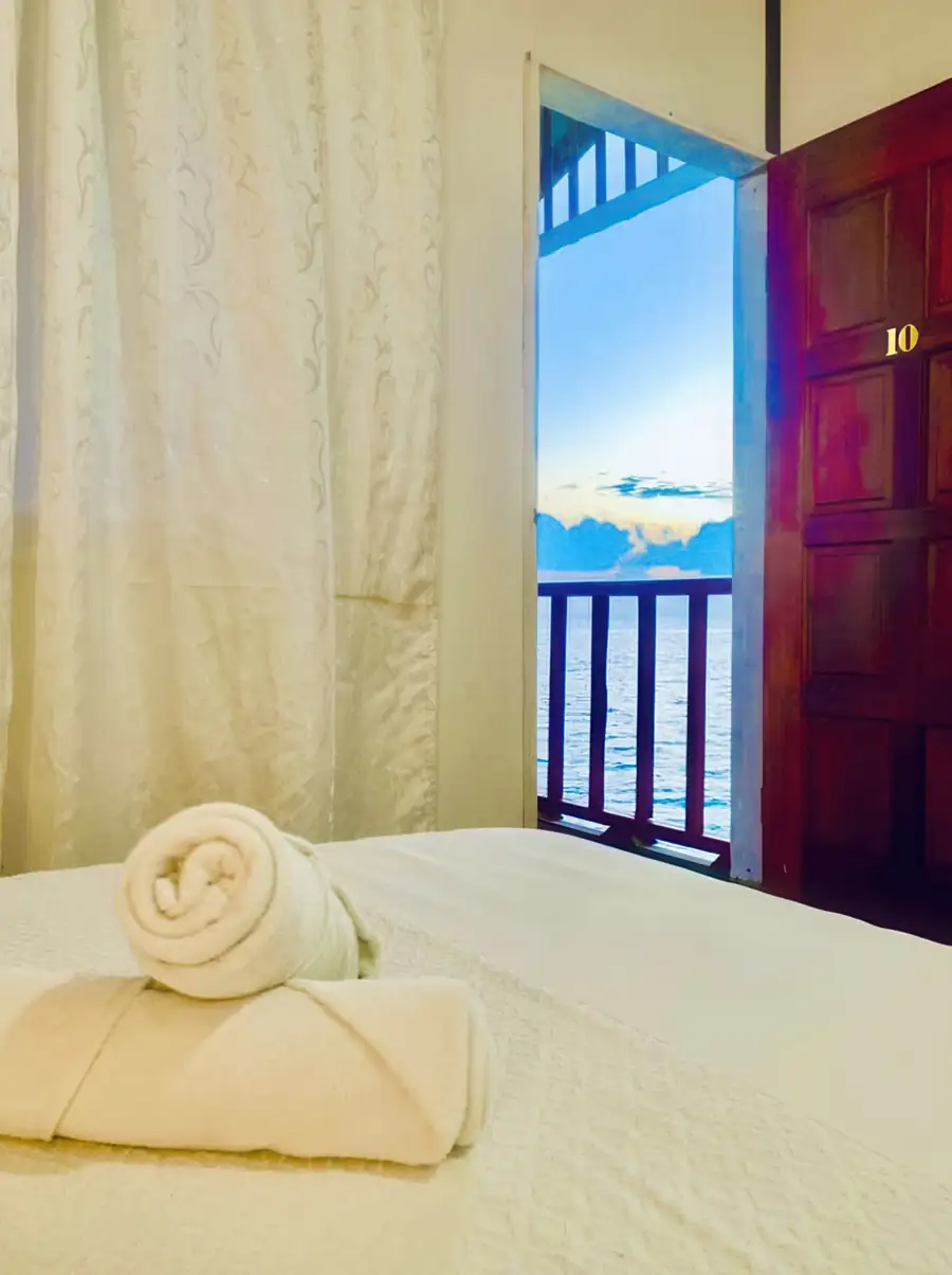 A tranquil bedroom at Bihing Angan Water Chalet highlighting a cozy bed, white towels, an open door leading to the balcony with ocean views.