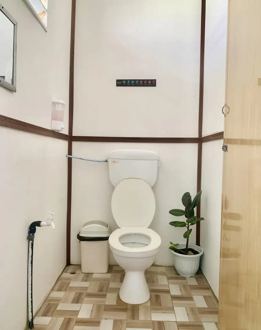 A rustic bathroom at Bihing Angan Water Chalet detailing charming wood-paneled walls, a classic washbasin, wall mirror, white toilet, and electric shower.