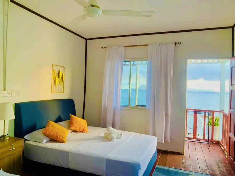 An inviting bedroom at Bihing Angan Water Chalet featuring a large bed, modern style bedside table, smooth wooden floors, A/C, and an open door to the oceanfront balcony.
